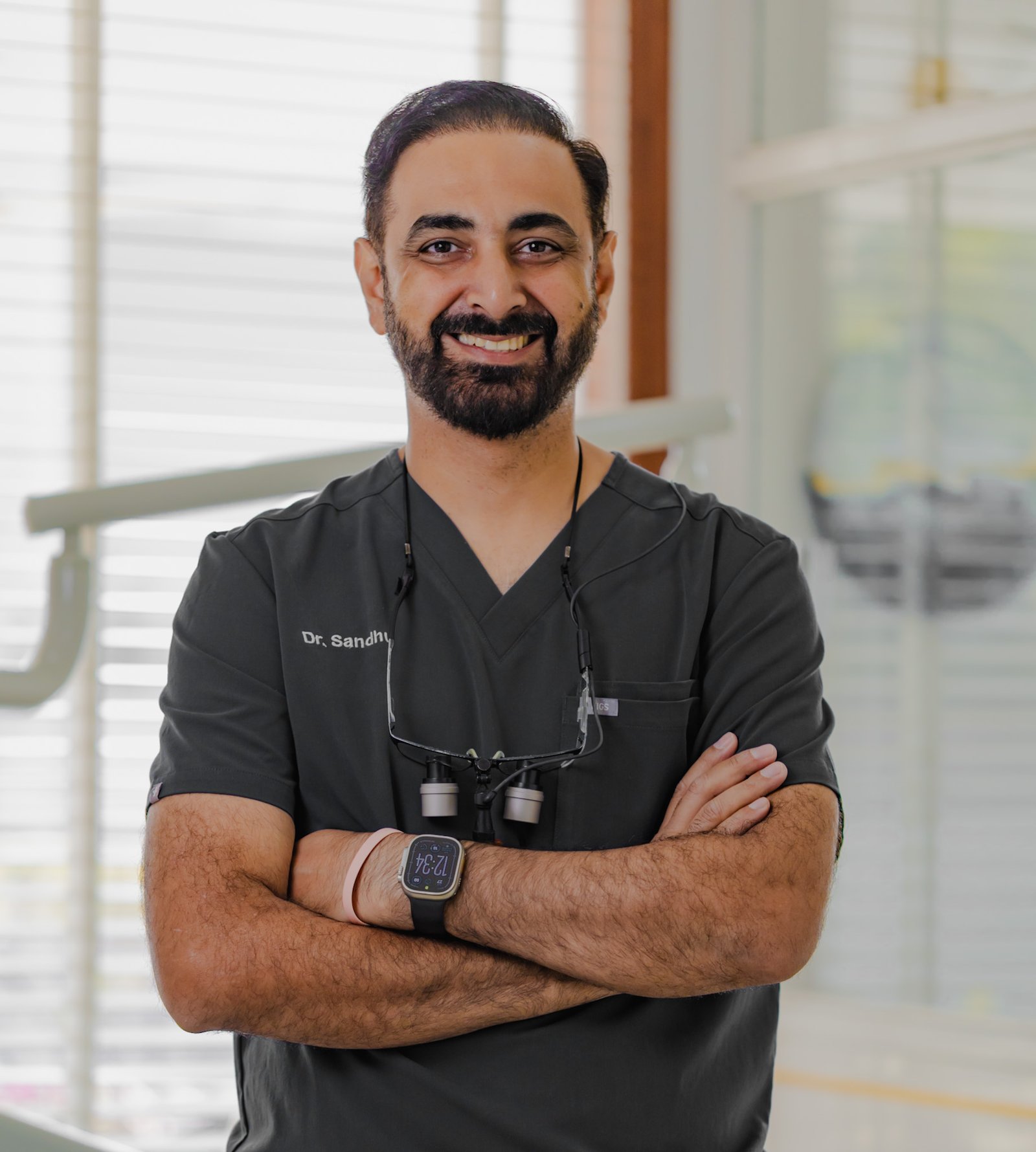 Dr. Sandhu, arms crossed wearing loupes in the clinic